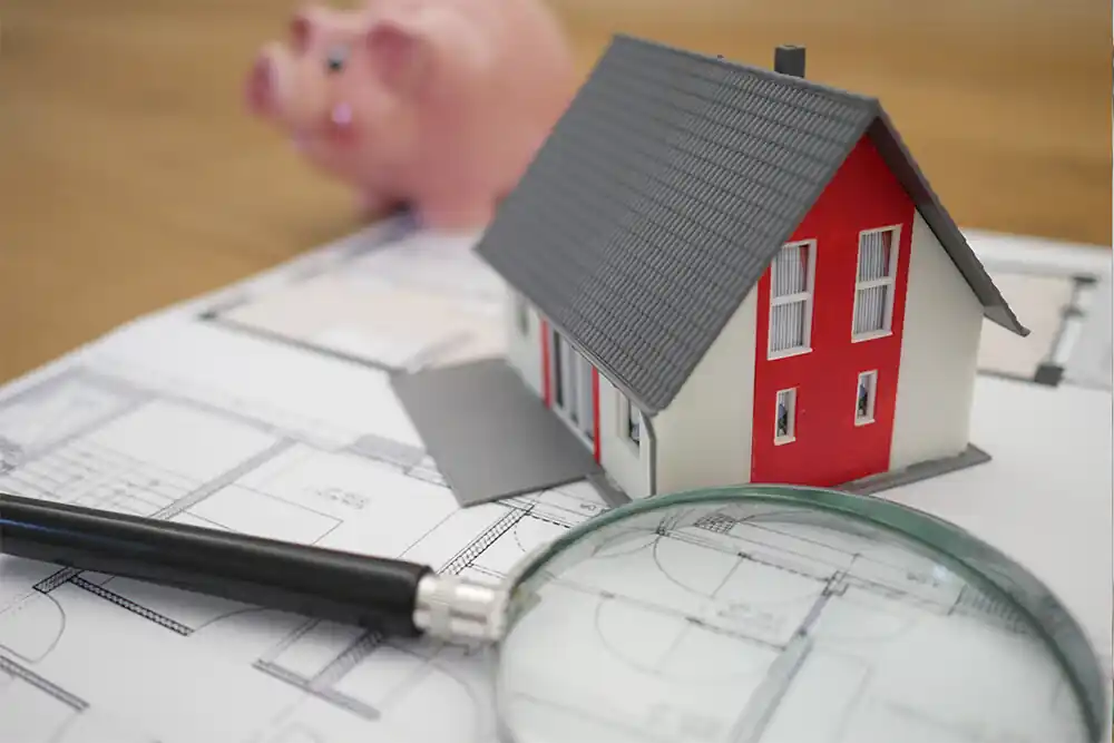 Property prices in Brisbane 2024 Property prices Brisbane with a magnifying glass in the foreground, a house model and pink piggy bank on top of a house plan.